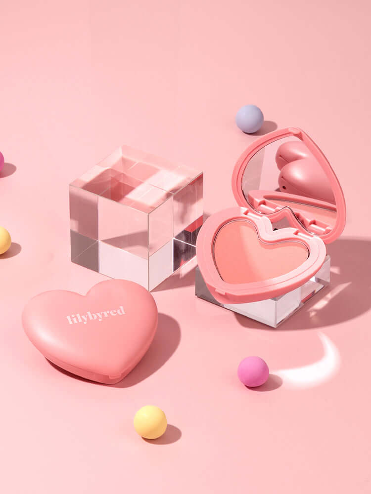 LILYBYRED Luv Beam Cheek 4.7g highlighter in heart-shaped compact on pink background with colorful balls.