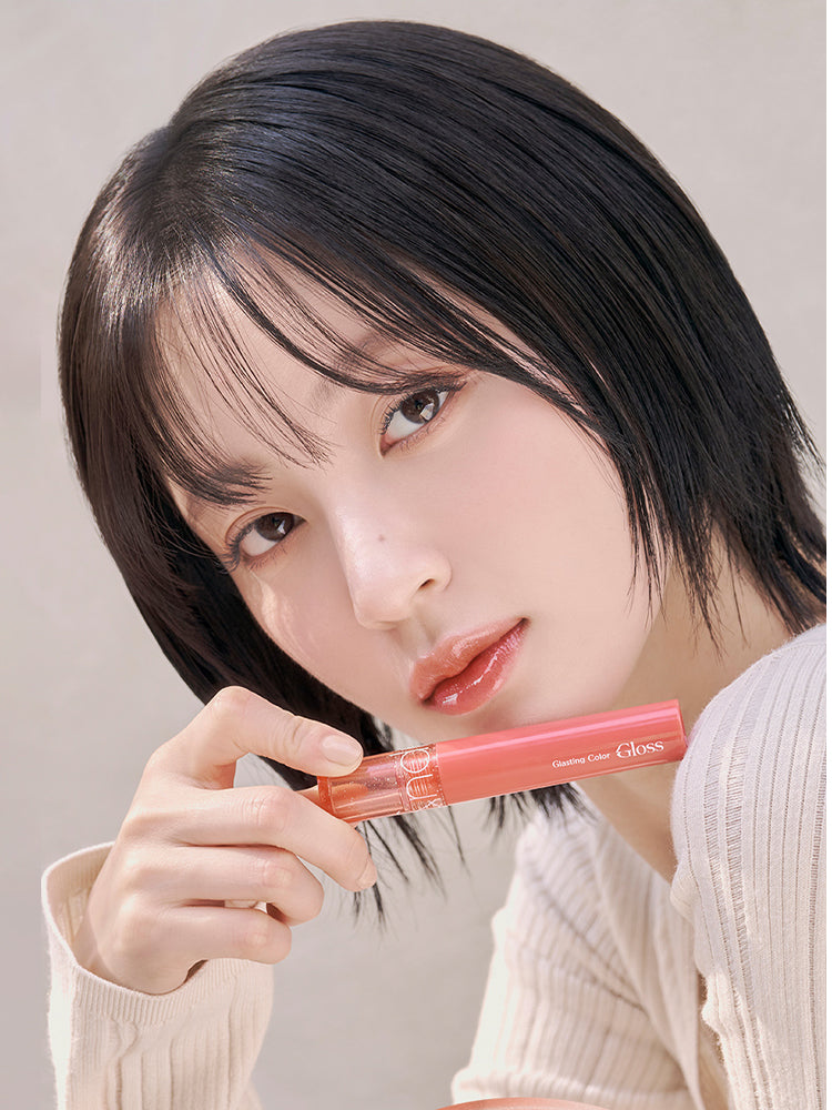 rom&nd Glasting Color Gloss Peach Sparkle showcased by a model holding the gloss, featuring a soft glow on her lips.