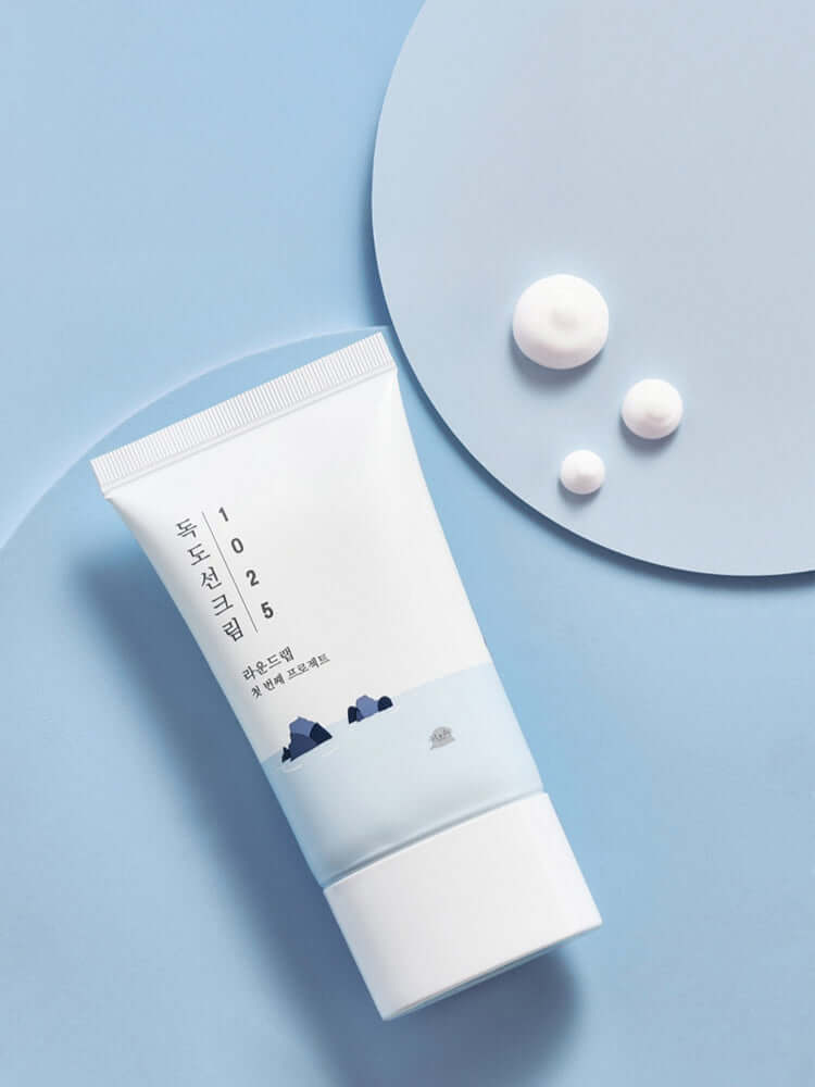 ROUND LAB 1025 Dokdo Sunscreen 50ml on light blue background with white cream drops.